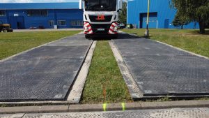 Droga wjazdowa z metalowymi płytami na trawie, z zaparkowanym pojazdem ciężarowym w tle.