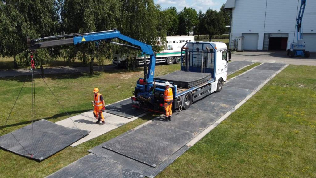 Czerwony samochód ciężarowy z podnośnikiem, dwóch pracowników w kamizelkach odblaskowych rozładowuje materiały na placu.