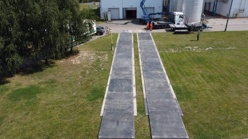 Dwie równoległe rampy z tworzywa sztucznego na trawie, obok pojazdy oraz pracownicy na placu przemysłowym.