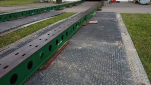 Długie zielone belki metalowe umieszczone na czarnej, teksturowanej nawierzchni na terenie budowy.