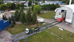 Ciężarówka z dźwigiem rozładunkowym, transportująca materiały przed dużym budynkiem przemysłowym, z otaczającą zielenią.