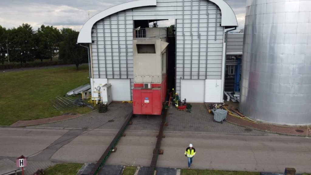 Wjazd dużego pojazdu do budynku przemysłowego, pracownicy w kamizelkach odblaskowych w tle.