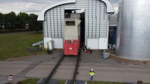 Wjazd dużego pojazdu do budynku przemysłowego, pracownicy w kamizelkach odblaskowych w tle.