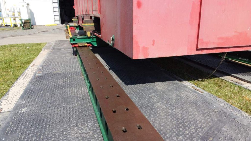 Czerwony kontener umieszczony na stalowej płycie, ustawiony na zielonej platformie na ziemi.