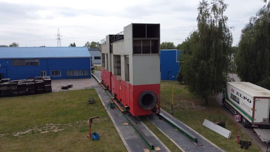 Duża konstrukcja przypominająca kontener, stojąca na wózkach, w otoczeniu budynków przemysłowych i zieleni.