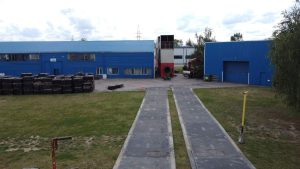 Zakład przemysłowy z niebieską fasadą, zewnętrzna waga samochodowa oraz składowane materiały.