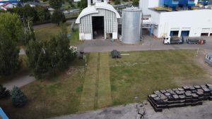 Widok na zakład przemysłowy z dużym budynkiem, silosem, terenami zielonymi oraz materiałami składowymi.