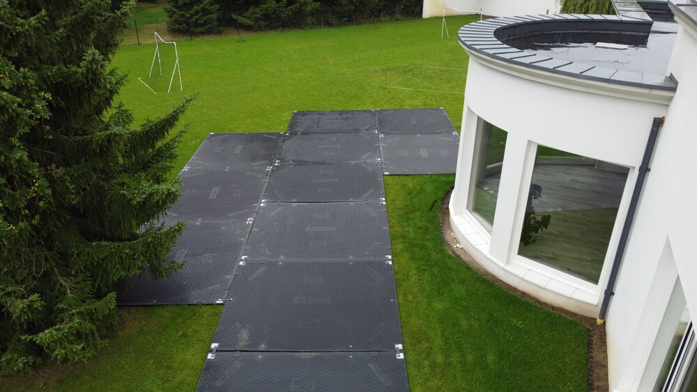 Czarna nawierzchnia z płytami na zielonej trawie prowadzi do białego budynku z dużymi oknami. W tle drzewa i huśtawka.