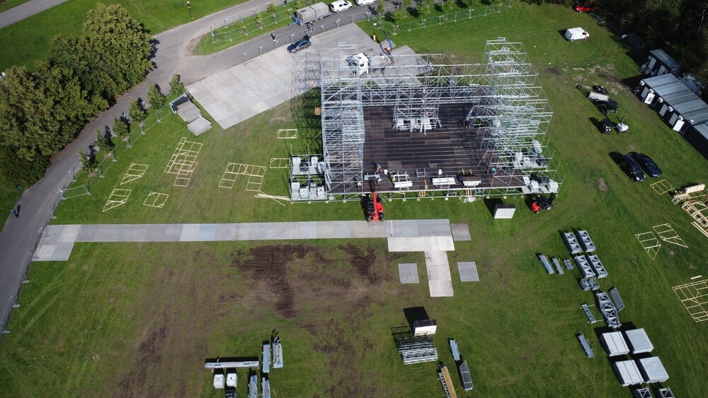 Widok z góry na plac budowy, gdzie znajduje się stalowa konstrukcja, maszyny oraz równo wykoszona trawa.