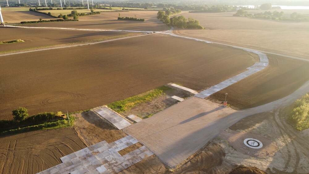 Widok na pola uprawne z utwardzoną nawierzchnią i fragmentem drogi w tle, otoczone zielenią drzew.
