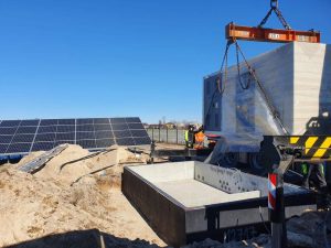 W budowie instalacja fotowoltaiczna, dźwig podnosi prefabrykat w otoczeniu piasku i wykopu.