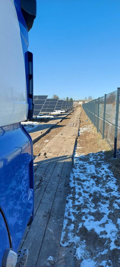 Widok na panele słoneczne z boiskiem, obok niebieski pojazd i śnieg na ziemi. Bezchmurne niebo.