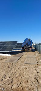 Maszyna z niebieską ciężarówką pracuje przy panelach słonecznych na piaszczystym terenie pod błękitnym niebem.
