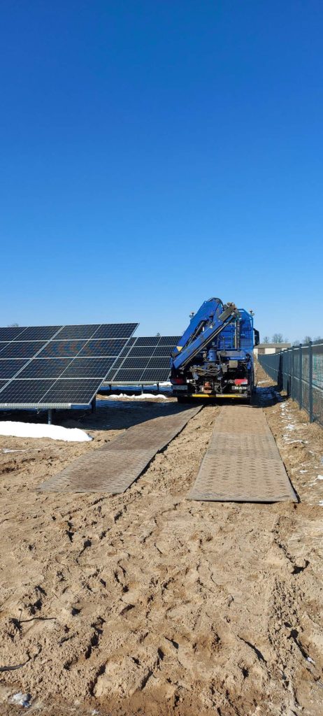 Maszyna z niebieską ciężarówką pracuje przy panelach słonecznych na piaszczystym terenie pod błękitnym niebem.
