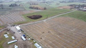 Widok z lotu ptaka na pola uprawne z wytyczonymi rzędami i sprzętem budowlanym w tle.