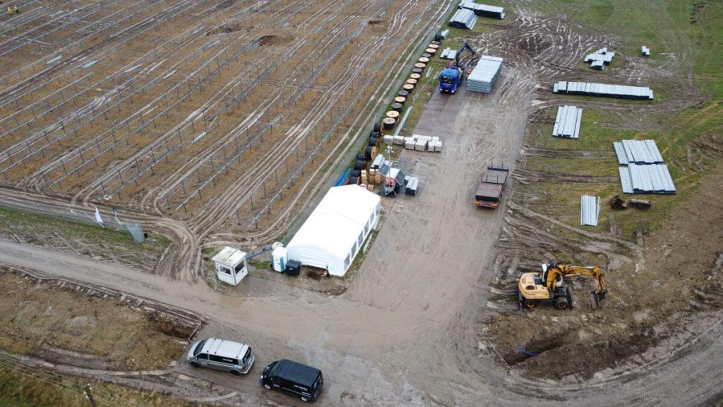 Widok na teren budowy z namiotem, maszynami, materiałami budowlanymi i śladami pojazdów na błotnistym podłożu.