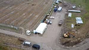Widok na teren budowy z namiotem, maszynami, materiałami budowlanymi i śladami pojazdów na błotnistym podłożu.