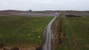 Widok na otwarte tereny, pokryte zieloną trawą, z widoczną drogą oraz liniami elektrycznymi. W tle widać strefy upraw.