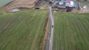 Widok z lotu ptaka na wiejską drogę, pola, zabudowania oraz pojazdy na drodze.