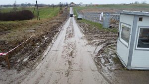 Droga pokryta błotem z wyraźnymi śladami kół, po obu stronach pola oraz budka ochrony. W tle widać pola i zagrody.