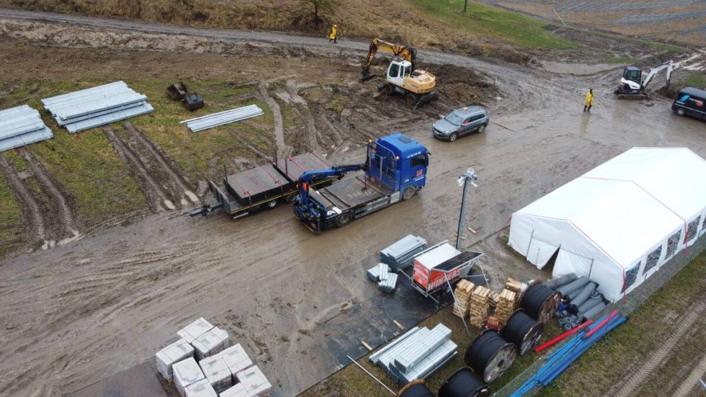Na obrazie widać plac budowy z błotem, ciężarówki, sprzęt budowlany oraz namiot. W tle widać wzniesienia.