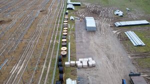 Widok na teren budowy z metalowymi stelażami, wałkami kabli, paczkami materiałów i śladami po sprzęcie w błocie.