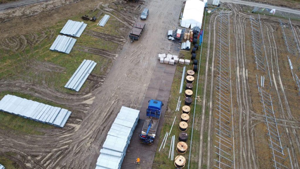 Widok z lotu ptaka na plac budowy z materiałami, pojazdami i prowizorycznymi halami. W tle stoją słupy.