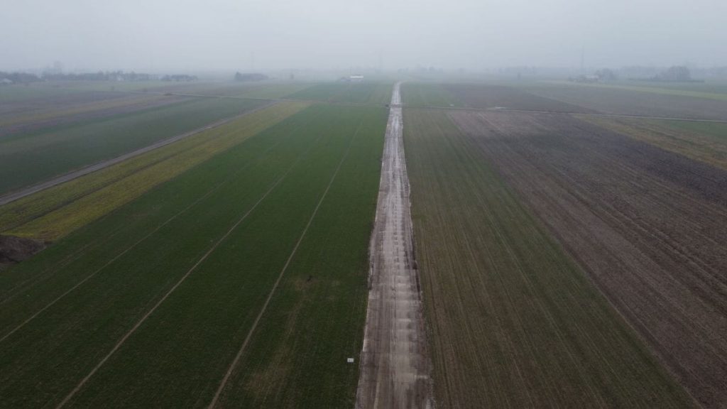 Widok na rozległe pola, z wyraźną drogą w centrum i mglistym niebem. W tle widać zabudowania.