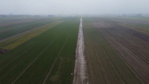 Widok na rozległe pola, z wyraźną drogą w centrum i mglistym niebem. W tle widać zabudowania.