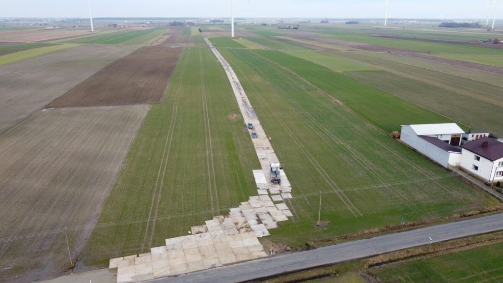Widok z lotu ptaka na wiejskie tereny z betonową drogą, ciągnącą się przez pola uprawne. W tle widoczne wiatraki.
