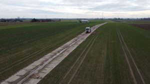 Ciężarówka na wiejskiej drodze otoczonej polami, w tle domy i szare, pochmurne niebo.