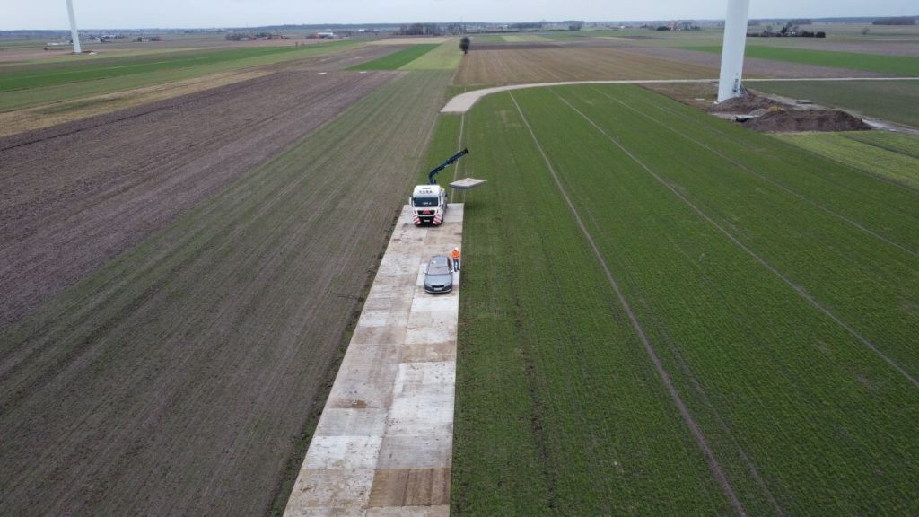Droga asfaltowa otoczona zielonymi polami, na której stoi samochód oraz urządzenie budowlane. W tle widać maszyny wiatrowe.