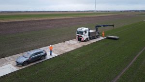 Pracownicy w pomarańczowych strojach na polu, obok ciężarówki i samochodu osobowego na betonowej nawierzchni.