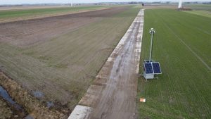 Widok na pole z panelami solarnymi oraz ścieżką, zarośniętymi brzegami i niewielkim rowem melioracyjnym.