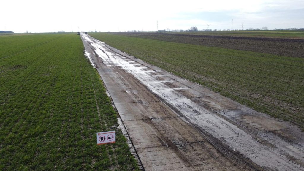 Droga z oznakowaniem ograniczenia prędkości, otoczona zielonymi polami, mokra nawierzchnia po deszczu.