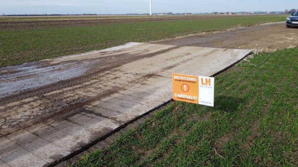 Widok na pole z utwardzoną drogą i znakiem ostrzegawczym o treści OSTRZEŻENIE! i CAREFULLY! w tle.