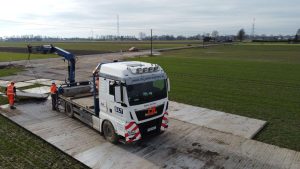 Ciężarówka z dźwigiem na polu, obok pracownicy w pomarańczowych strojach, otoczona zielonymi uprawami.