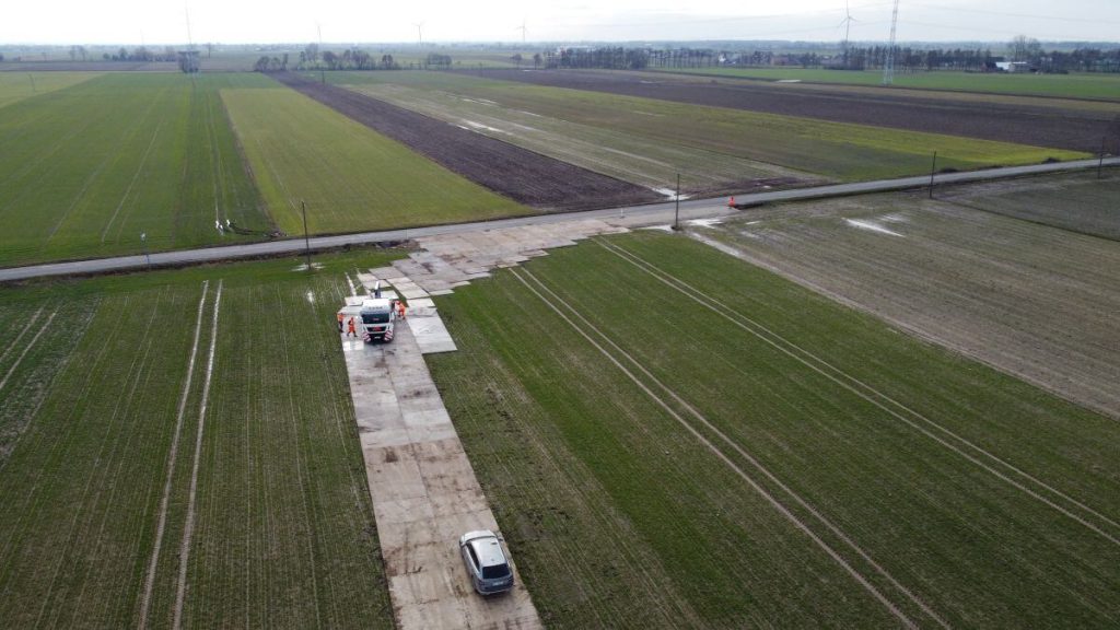 Widok na pola uprawne z drogą i zaparkowanym samochodem. W tle widoczne elektrownie wiatrowe.