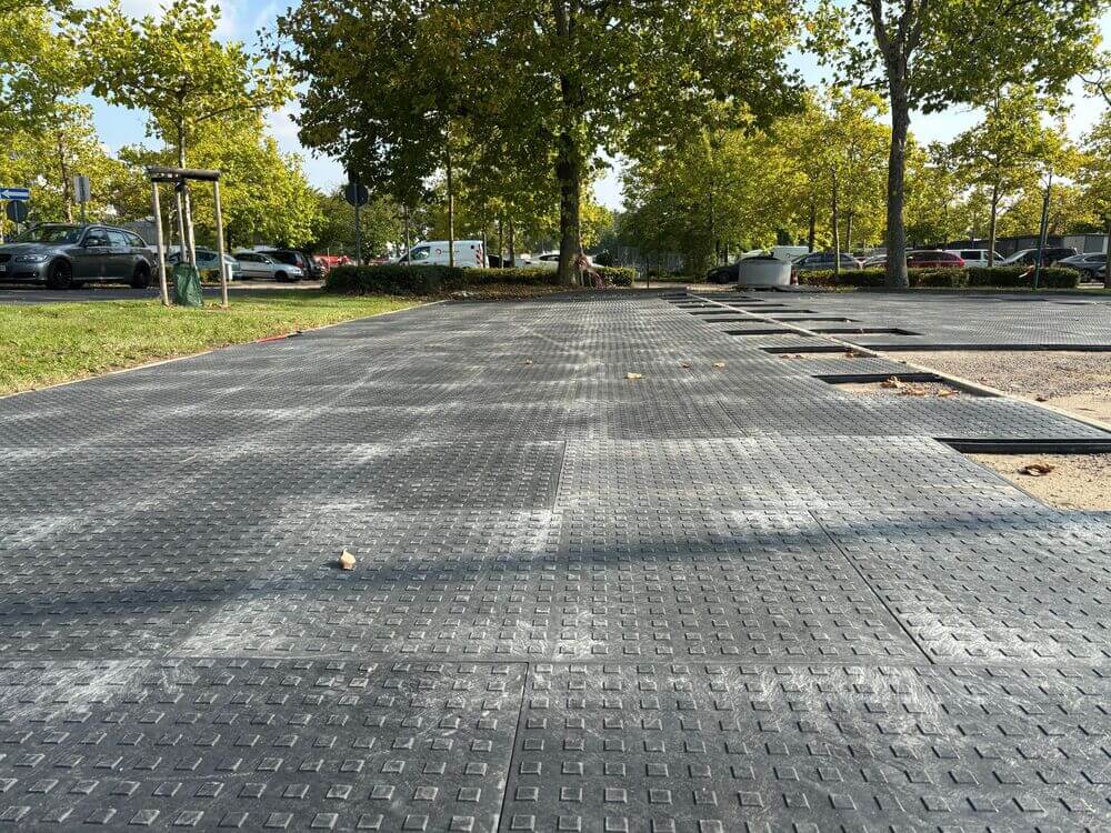 Widok na czarne płyty na parkingu, z drzewami w tle i częściowo odsłoniętymi miejscami na ziemię.