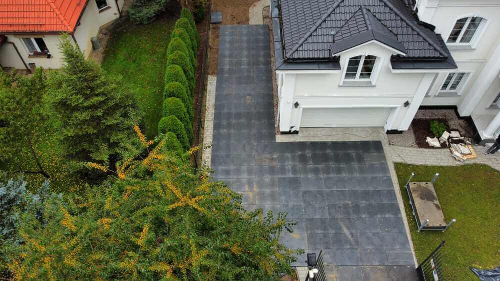 Widok z góry na podjazd do nowoczesnego domu, obłożony szarym kostką, otoczony zielonymi krzewami.