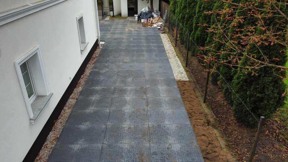 Podjazd wyłożony szaro-czarnymi kostkami, otoczony zaroślami i ścianą budynku. W tle leży sterta odpadów.