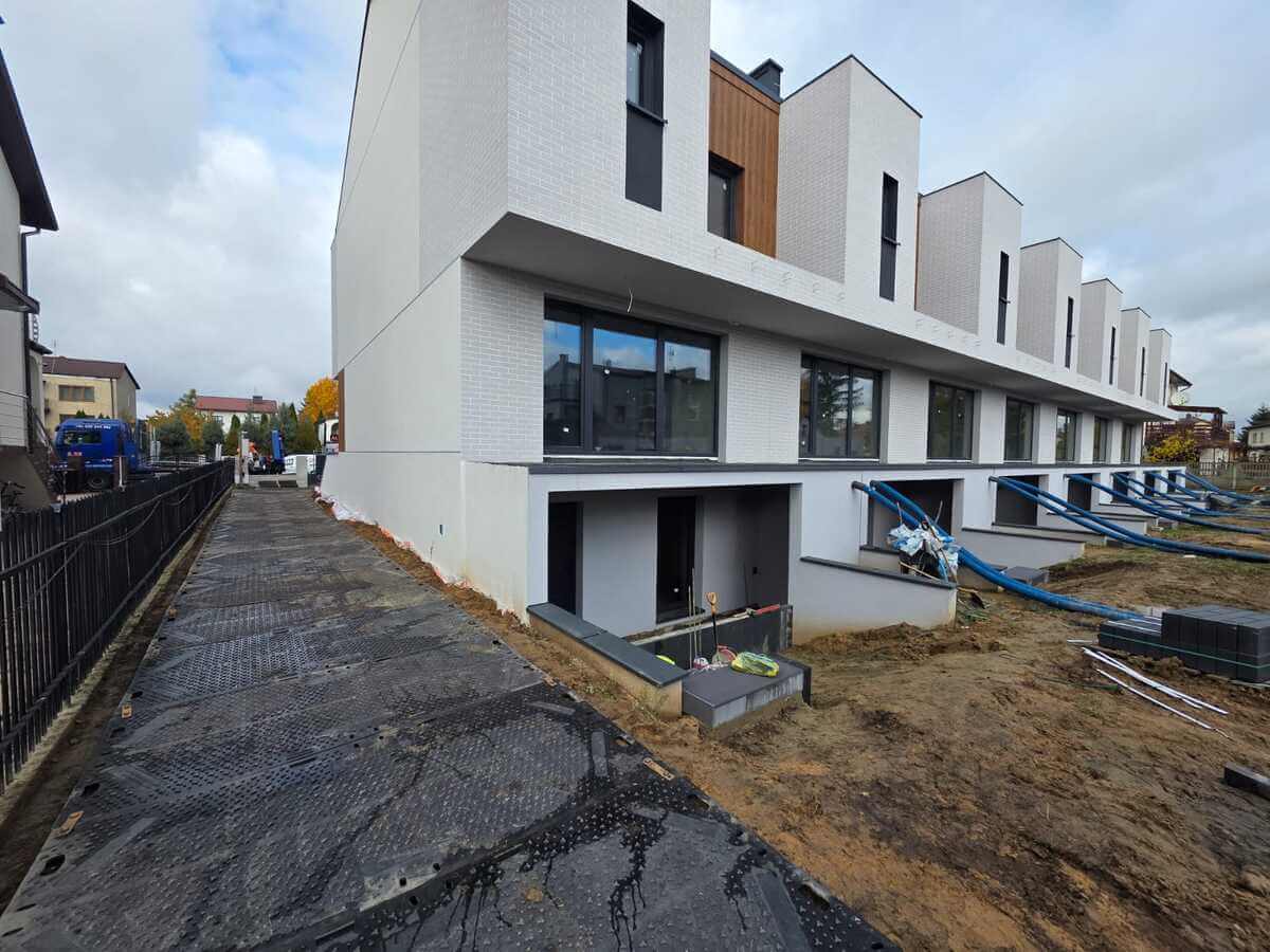 Nowoczesny budynek mieszkalny z białą elewacją, otoczony placem budowy oraz wyłożonym chodnikiem.