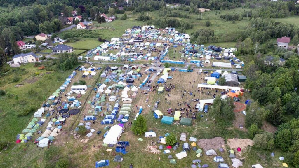 Widok z lotu ptaka na obozowisko z namiotami w zielonym terenie, otoczonym lasami i zabudową wiejską.