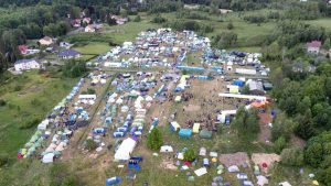 Widok z lotu ptaka na obozowisko z namiotami w zielonym terenie, otoczonym lasami i zabudową wiejską.