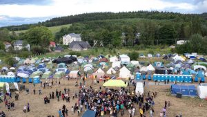 Obozowisko z licznymi namiotami, grupą ludzi oraz toaletami przenośnymi, otoczone zieleń i budynki.