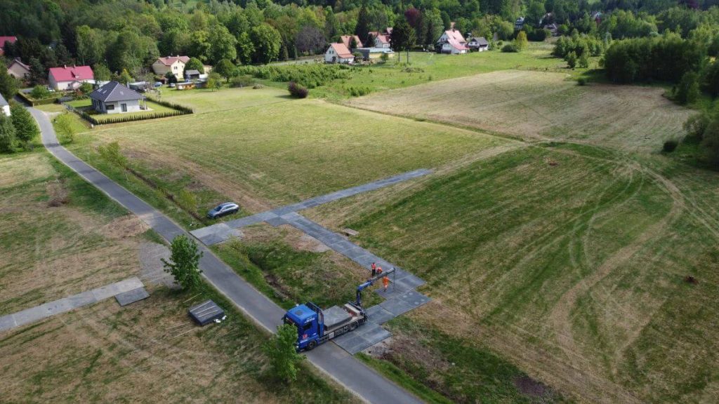 Widok z lotu ptaka na wiejski krajobraz z drogą, pojazdem budowlanym i domami na tle zielonych pól.
