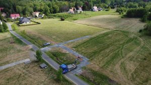 Widok z lotu ptaka na wiejski krajobraz z drogą, pojazdem budowlanym i domami na tle zielonych pól.