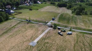 Widok z góry na działkę z drogą, kilkoma pojazdami i budynkami w tle, otoczoną zielonymi terenami.