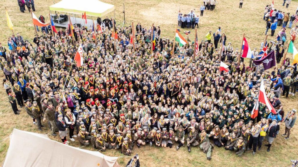 Grupa skautów w mundurach, trzymających flagi, pozująca do zdjęcia na terenie wydarzenia na świeżym powietrzu.