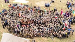 Grupa skautów w mundurach, trzymających flagi, pozująca do zdjęcia na terenie wydarzenia na świeżym powietrzu.